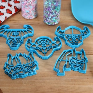 May include: Five blue plastic cookie cutters shaped like cartoon monster faces with horns. The cutters are on a wooden surface with two jars of sprinkles and a blue spatula.