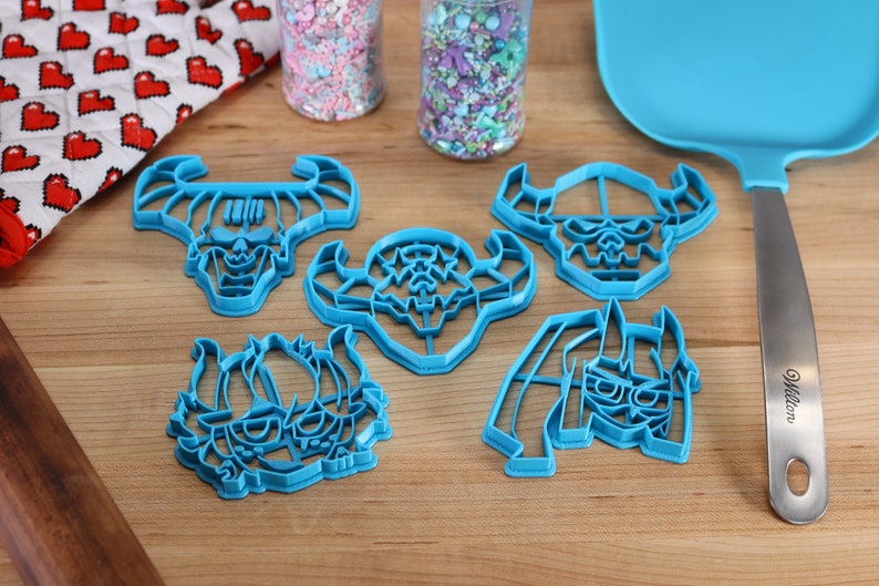 May include: Five blue plastic cookie cutters shaped like cartoon monster faces with horns. The cutters are on a wooden surface with two jars of sprinkles and a blue spatula.