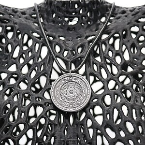 May include: A silver pendant necklace with a circular pendant featuring a detailed geometric design. The necklace is displayed on a black, textured mannequin.