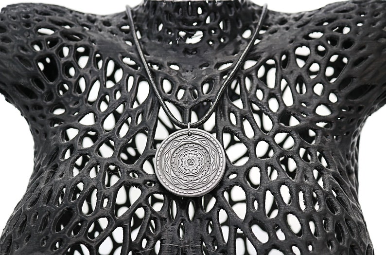 May include: A silver pendant necklace with a circular pendant featuring a detailed geometric design. The necklace is displayed on a black, textured mannequin.