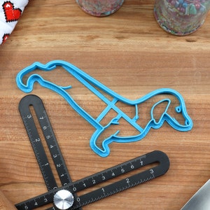 May include: A blue plastic cookie cutter shaped like a dachshund dog. The cutter is on a wooden surface with a ruler and a rolling pin.