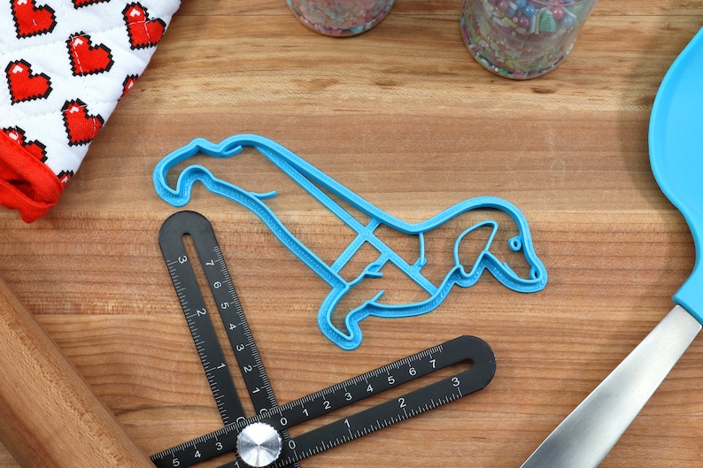 May include: A blue plastic cookie cutter shaped like a dachshund dog. The cutter is on a wooden surface with a ruler and a rolling pin.