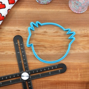 May include: A blue plastic cookie cutter shaped like a bird's head on a wooden surface with a black measuring tool.