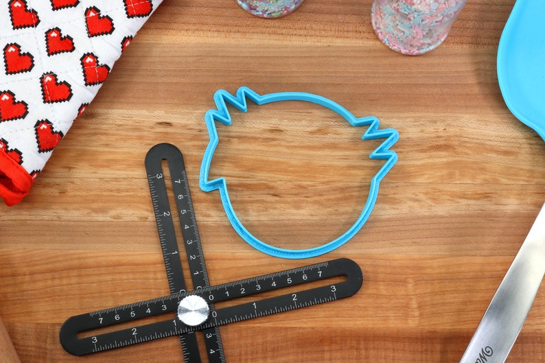 May include: A blue plastic cookie cutter shaped like a bird's head on a wooden surface with a black measuring tool.