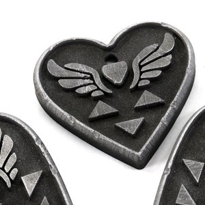 Puede incluir: Tres dijes de coraz&oacute;n plateados con un s&iacute;mbolo alado y un dise&ntilde;o triangular.