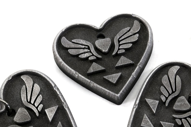 Puede incluir: Tres dijes de coraz&oacute;n plateados con un s&iacute;mbolo alado y un dise&ntilde;o triangular.