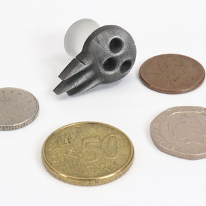 May include: A dark gray metal bottle stopper with a unique design featuring three circular holes and two wing-like protrusions. Several coins of different sizes and colors are arranged around the stopper on a white surface.