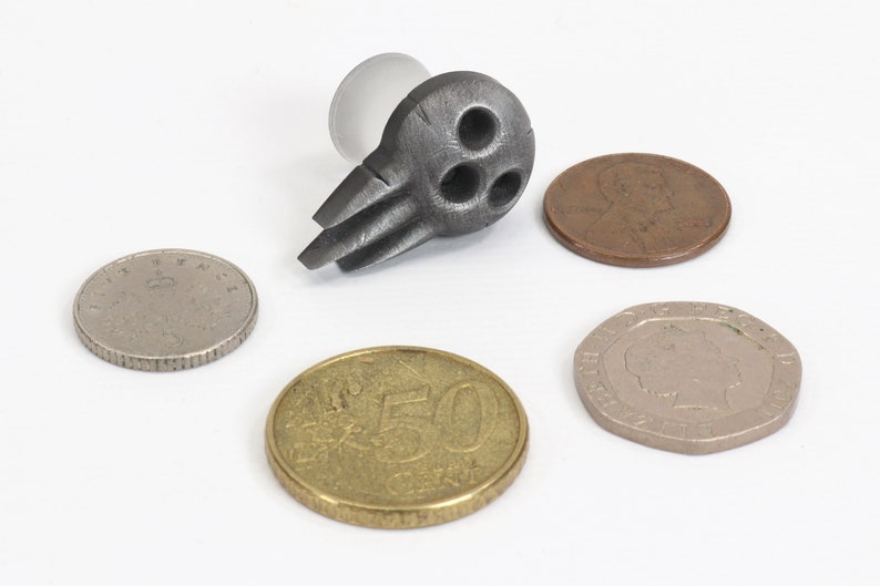 May include: A dark gray metal bottle stopper with a unique design featuring three circular holes and two wing-like protrusions. Several coins of different sizes and colors are arranged around the stopper on a white surface.