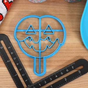 May include: Blue plastic cookie cutter shaped like a jack-o'-lantern with a dripping face. The cutter has a stick handle.