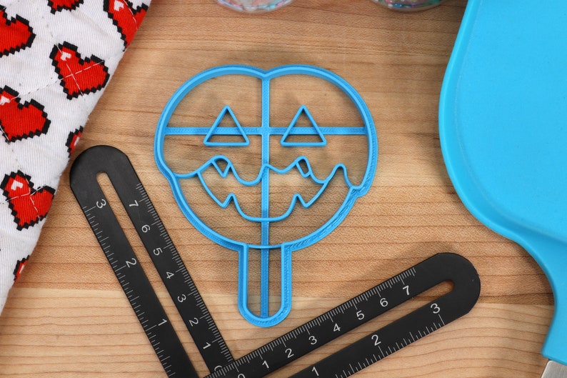 May include: Blue plastic cookie cutter shaped like a jack-o'-lantern with a dripping face. The cutter has a stick handle.