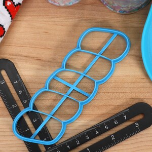 May include: A blue plastic cookie cutter shaped like a row of five connected circles. The cutter is on a wooden surface with black rulers.