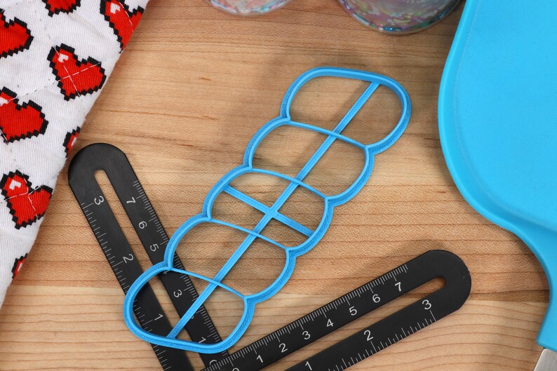 May include: A blue plastic cookie cutter shaped like a row of five connected circles. The cutter is on a wooden surface with black rulers.