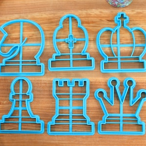 May include: Six blue plastic cookie cutters shaped like chess pieces: a knight, a bishop, a king, a pawn, a rook, and a queen.