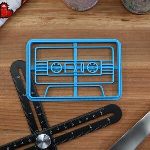 May include: A blue plastic cookie cutter shaped like a cassette tape. The cookie cutter is on a wooden surface with a ruler and a blue spatula.