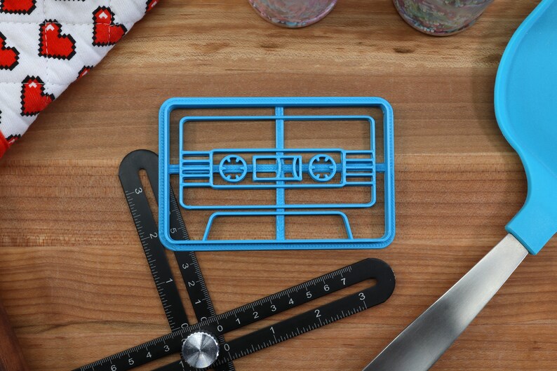 May include: A blue plastic cookie cutter shaped like a cassette tape. The cookie cutter is on a wooden surface with a ruler and a blue spatula.