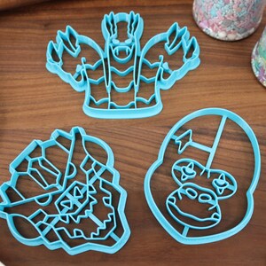 May include: Three blue plastic cookie cutters shaped like cartoon monster faces. The cutters are on a wooden surface.