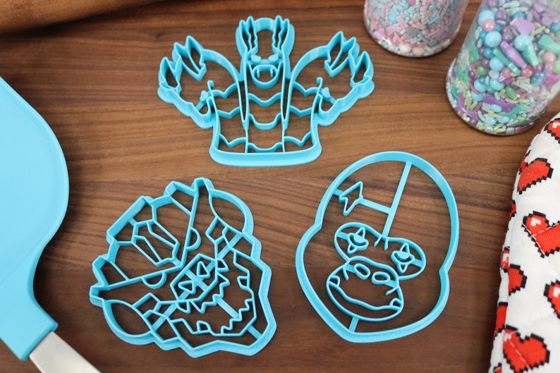 May include: Three blue plastic cookie cutters shaped like cartoon monster faces. The cutters are on a wooden surface.