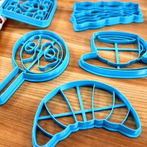 May include: A collection of bright blue cookie cutters in various shapes, including a lollipop, a hat, and a rectangular shape with a grid pattern. The cutters are arranged on a wooden surface.