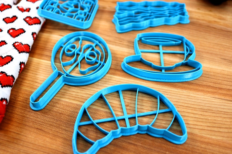 May include: A collection of bright blue cookie cutters in various shapes, including a lollipop, a hat, and a rectangular shape with a grid pattern. The cutters are arranged on a wooden surface.