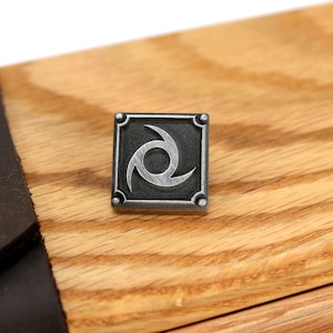 May include: A silver metal pin with a black background and a stylised silver swirl design. The pin is square shaped and has a raised border.
