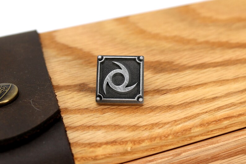 May include: A silver metal pin with a black background and a stylised silver swirl design. The pin is square shaped and has a raised border.