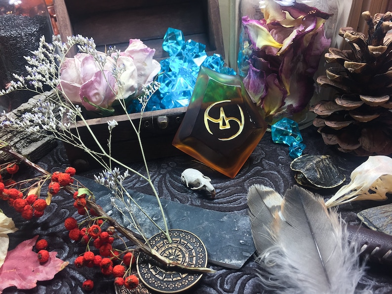 May include: A close-up of a table with a variety of objects, including a small wooden box with dried roses, a clear glass jar with dried rose petals, a pine cone, a small turtle figurine, a bird skull, a feather, a small stone with a symbol, and a small metal disc with a design.