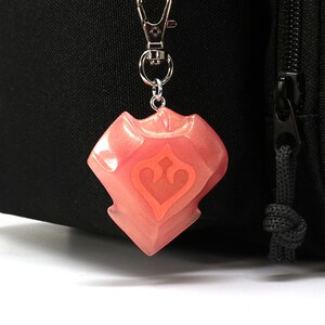 May include: A pink resin keychain shaped like a shield with a stylized heart design. The keychain is attached to a silver metal clasp.