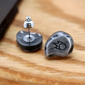 May include: A pair of stud earrings with a silver finish. Each earring has a rounded, stone-like shape with a black symbol. The earrings are displayed on a wooden surface.