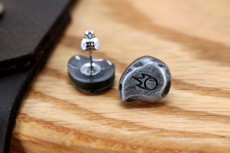 May include: A pair of stud earrings with a silver finish. Each earring has a rounded, stone-like shape with a black symbol. The earrings are displayed on a wooden surface.