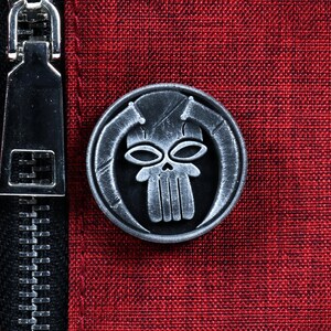 May include: A silver-colored metal pin with a skull design. The skull has two eye sockets and a set of teeth. The skull is surrounded by a circular frame.