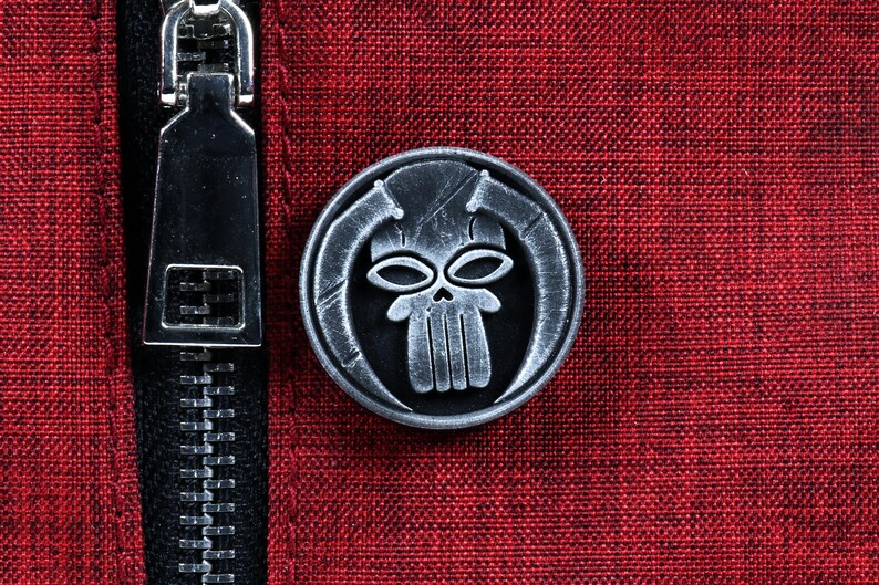 May include: A silver-colored metal pin with a skull design. The skull has two eye sockets and a set of teeth. The skull is surrounded by a circular frame.