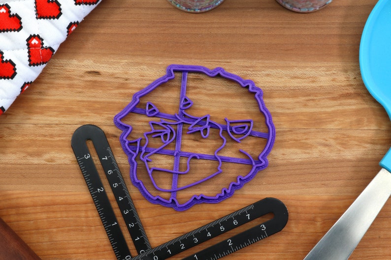 May include: Purple plastic cookie cutter shaped like a dog's head. The cutter has a detailed design with a fluffy texture.