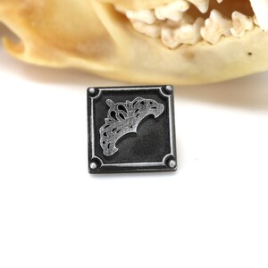 May include: Square, dark gray metal pendant with a decorative, stylized design. The pendant has rounded corners and a raised border. The design appears to be a stylized floral or organic shape. The background is white.