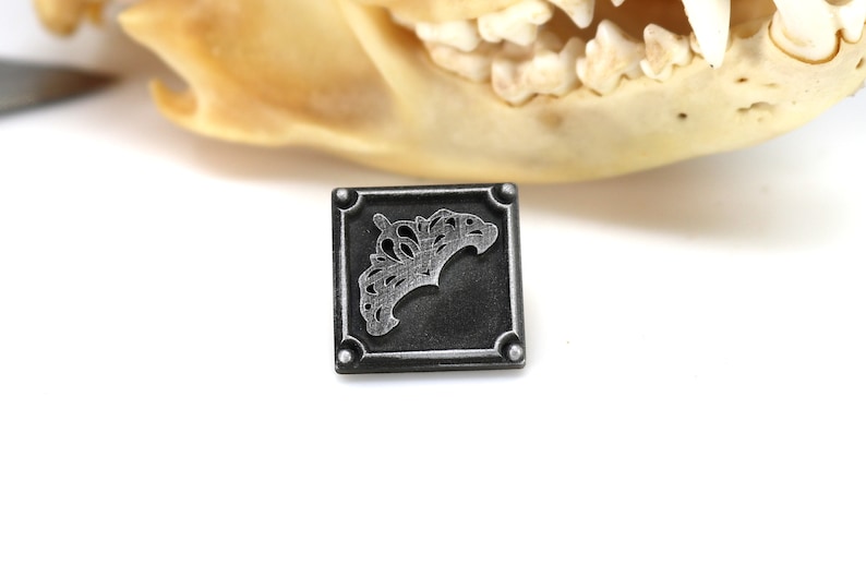 May include: Square, dark gray metal pendant with a decorative, stylized design. The pendant has rounded corners and a raised border. The design appears to be a stylized floral or organic shape. The background is white.