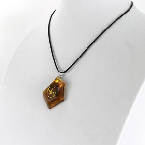 May include: A brown, faceted pendant with a gold symbol hangs from a black cord necklace. The pendant is shaped like a diamond.