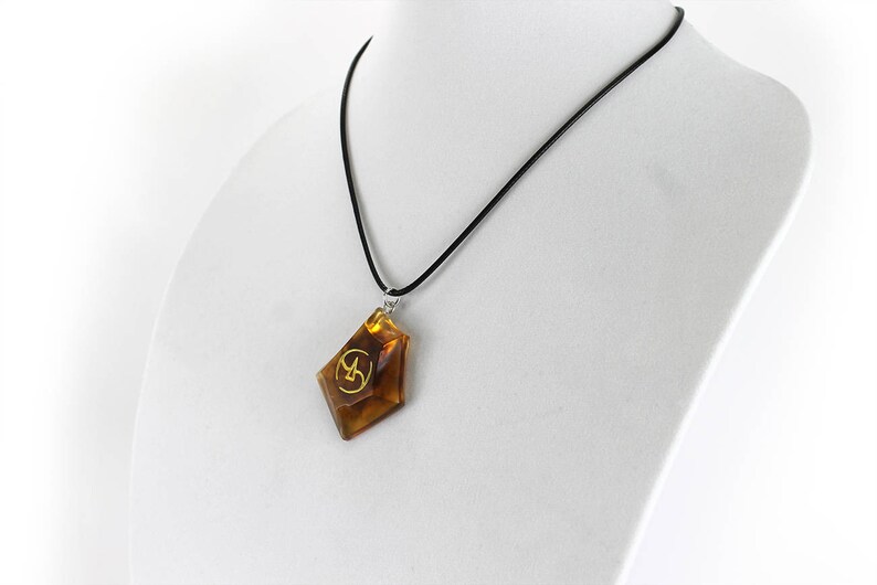 May include: A brown, faceted pendant with a gold symbol hangs from a black cord necklace. The pendant is shaped like a diamond.