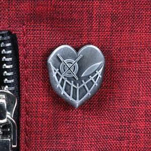 K&ouml;nnte beinhalten: Eine silberne herzf&ouml;rmige Anstecknadel mit einem gestickten Design und einem Kreuzaugen-Symbol. Die Anstecknadel ist auf einem roten Stoffhintergrund befestigt.