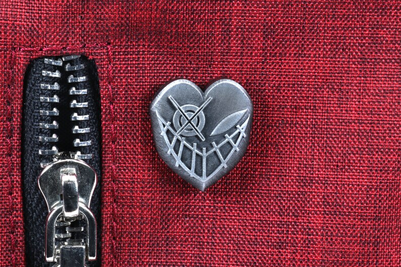 K&ouml;nnte beinhalten: Eine silberne herzf&ouml;rmige Anstecknadel mit einem gestickten Design und einem Kreuzaugen-Symbol. Die Anstecknadel ist auf einem roten Stoffhintergrund befestigt.