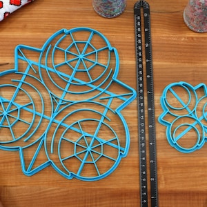 May include: Two blue plastic cookie cutters on a wooden surface. The larger cutter is shaped like a snowflake with a circular design. The smaller cutter is shaped like three overlapping circles.