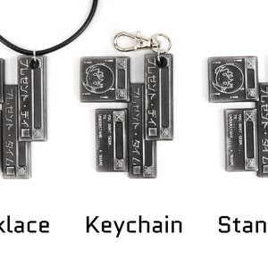 May include: A silver-toned metal pendant necklace, keychain, and standard charm featuring a stylized image of a character's face with the text "プレゼント・タイム" (present time) in Japanese. The charm is made up of five interlocking pieces.