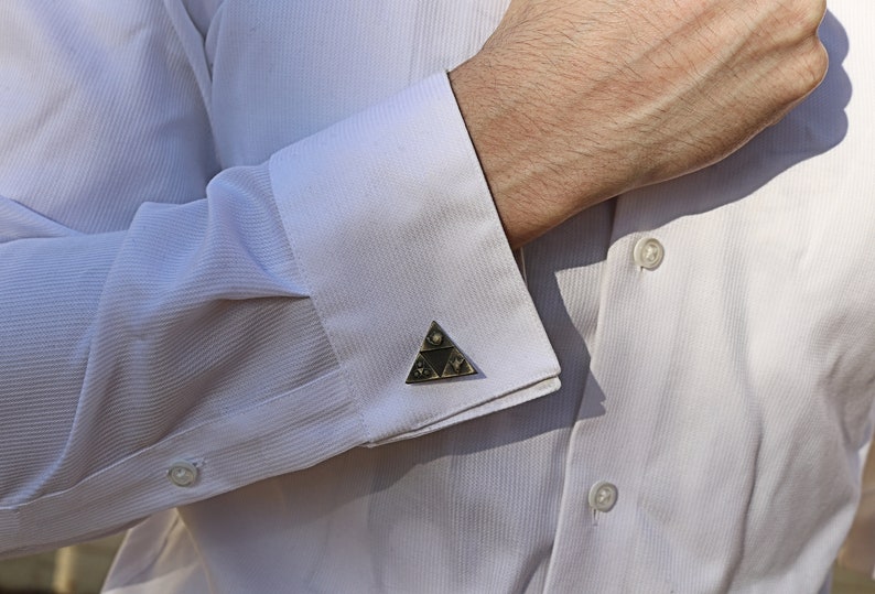 May include: A white dress shirt with a pair of cufflinks featuring a silver triangular design with black and white details.