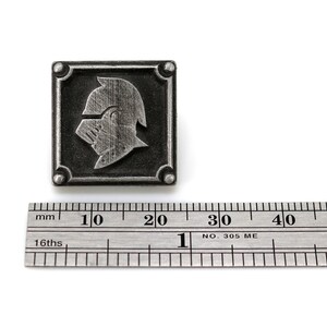 May include: A silver metal pin with a square design featuring a knight's helmet in profile. The pin is approximately 2 cm wide.