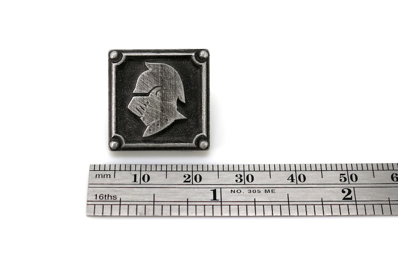 May include: A silver metal pin with a square design featuring a knight's helmet in profile. The pin is approximately 2 cm wide.
