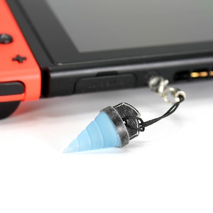 May include: A blue and silver drill bit charm attached to a black string. The charm is hanging from a Nintendo Switch console.