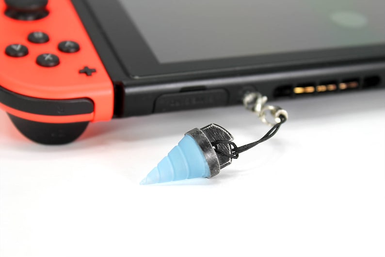 May include: A blue and silver drill bit charm attached to a black string. The charm is hanging from a Nintendo Switch console.