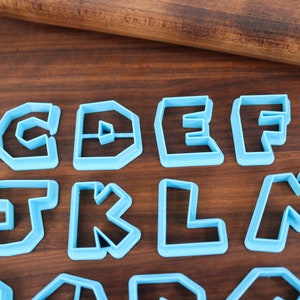 May include: A set of blue plastic cookie cutters shaped like the letters B, C, D, E, F, I, J, K, L, M, N, O, P, Q, R, S, T, U, V, W, X, Y, and Z. The cutters are arranged in three rows on a wooden surface.