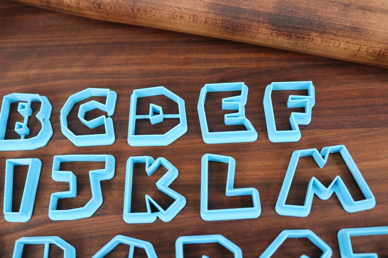 May include: A set of blue plastic cookie cutters shaped like the letters B, C, D, E, F, I, J, K, L, M, N, O, P, Q, R, S, T, U, V, W, X, Y, and Z. The cutters are arranged in three rows on a wooden surface.