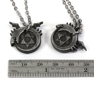 K&ouml;nnte beinhalten: Zwei silberne Halsketten mit Anh&auml;ngern, die das alchemistische Symbol f&uuml;r den Stein der Weisen zeigen, umgeben von einem Drachen-Design. Die Anh&auml;nger haben einen Durchmesser von etwa 2 cm.