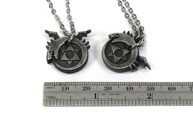 K&ouml;nnte beinhalten: Zwei silberne Halsketten mit Anh&auml;ngern, die das alchemistische Symbol f&uuml;r den Stein der Weisen zeigen, umgeben von einem Drachen-Design. Die Anh&auml;nger haben einen Durchmesser von etwa 2 cm.