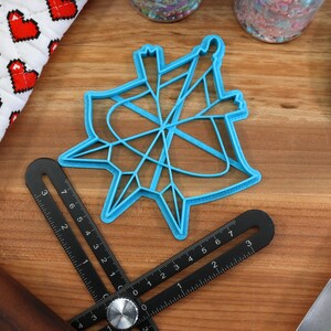 May include: A blue plastic cookie cutter shaped like a star with a geometric design. The cutter is on a wooden surface with a ruler, a rolling pin, and a spatula.